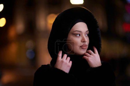 Téléchargez les photos : Femme musulmane marchant dans une rue urbaine par une froide nuit d'hiver portant le hijab avec des lumières de la ville bokeh en arrière-plan - en image libre de droit