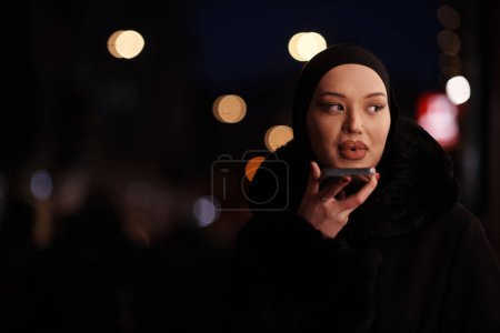 Téléchargez les photos : Belle et élégante européenne musulmane Hijabi Business Lady vérifier son téléphone et les médias sociaux sur la rue de la ville urbaine la nuit - en image libre de droit