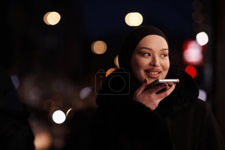 Photo for Beautiful and elegant European Muslim Hijabi Business Lady checking her phone and social media on urban city street at night - Royalty Free Image