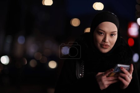 Foto de Hermosa y elegante musulmana europea Hijabi Business Lady revisando su teléfono y redes sociales en la calle urbana de la ciudad por la noche - Imagen libre de derechos