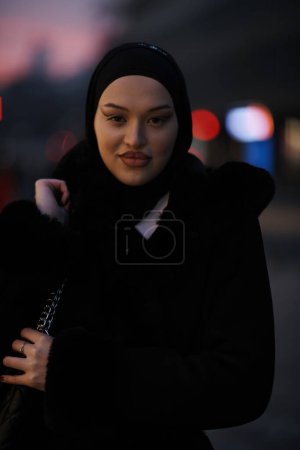 Téléchargez les photos : Femme musulmane marchant dans une rue urbaine par une froide nuit d'hiver portant le hijab avec des lumières de la ville bokeh en arrière-plan - en image libre de droit
