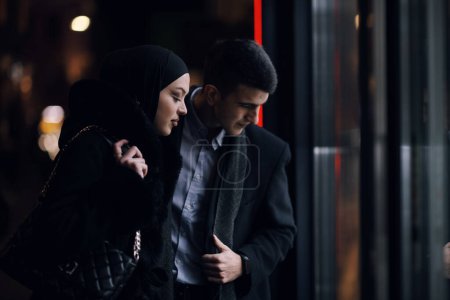 Foto de Feliz pareja de negocios multiculturales caminando juntos al aire libre en una calle urbana de la ciudad por la noche cerca de un escaparate de joyería. Exitoso empresario árabe y mujer musulmana europea. - Imagen libre de derechos