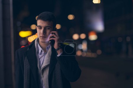 Photo for Smiling Meedle Eastern man walking down street near modern office building, freelancer businessman looking away holding mobile phone on busy city street at night - Royalty Free Image