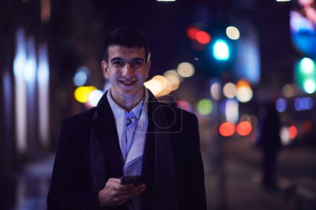 Téléchargez les photos : Souriant Meedle Eastern homme marchant dans la rue près de l'immeuble de bureaux moderne, homme d'affaires indépendant regardant loin tenant téléphone mobile sur la rue animée de la ville la nuit - en image libre de droit