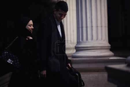 Foto de Feliz pareja de negocios multiculturales caminando juntos al aire libre en una calle urbana de la ciudad por la noche cerca de un escaparate de joyería. Exitoso empresario árabe y mujer musulmana europea. - Imagen libre de derechos