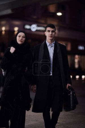 Photo for Happy multicultural business couple walking together outdoors in an urban city street at night near a jewelry shopping store window. Successful Arab businessman and European Muslim woman. - Royalty Free Image