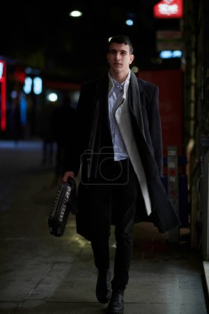 Photo for Smiling Meedle Eastern man walking down street near modern office building, freelancer businessman looking away holding mobile phone on busy city street at night - Royalty Free Image