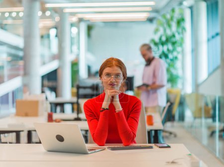 Téléchargez les photos : Dans un bureau de start-up moderne, une femme d'affaires professionnelle aux cheveux oranges assise à son ordinateur portable, incarnant l'innovation et la productivité dans son espace de travail contemporain. - en image libre de droit