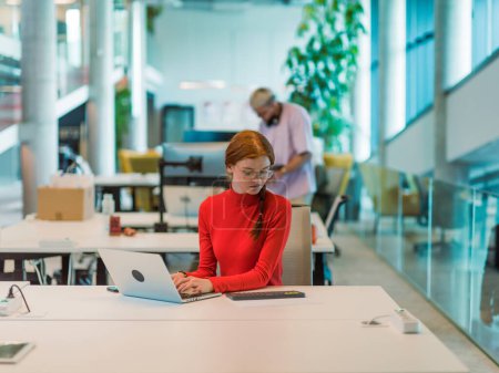Foto de En una moderna startup, una empresaria profesional con el pelo naranja sentada en su portátil, personificando la innovación y la productividad en su espacio de trabajo contemporáneo. - Imagen libre de derechos