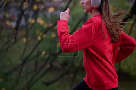 Téléchargez les photos : Mode de vie sain, jogging matinal. Jeune belle femme qui court dans le parc d'automne et écoute de la musique avec un casque sur smartphone. Porter une chemise de sport rouge. - en image libre de droit