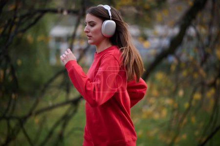 Photo for Healthy lifestyle, morning jogging. Young beautiful woman running in autumn park and listening to music with headphones on smartphone. Wearing a red sport shirt. - Royalty Free Image