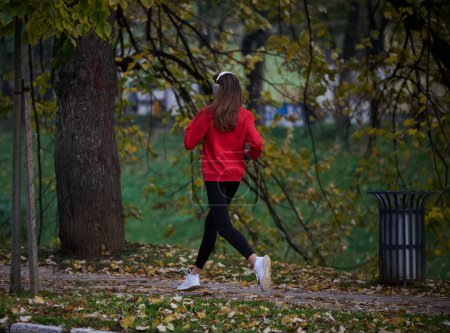 Foto de Un estilo de vida saludable, correr por la mañana. Mujer hermosa joven corriendo en el parque de otoño y escuchando música con auriculares en el teléfono inteligente. Usar una camisa deportiva roja. - Imagen libre de derechos