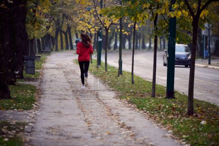 Téléchargez les photos : Mode de vie sain, jogging matinal. Jeune belle femme qui court dans le parc d'automne et écoute de la musique avec un casque sur smartphone. Porter une chemise de sport rouge. - en image libre de droit