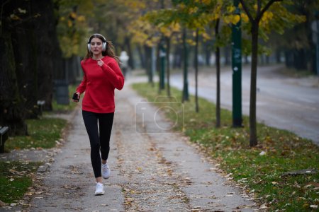 Photo for Healthy lifestyle, morning jogging. Young beautiful woman running in autumn park and listening to music with headphones on smartphone. Wearing a red sport shirt. - Royalty Free Image