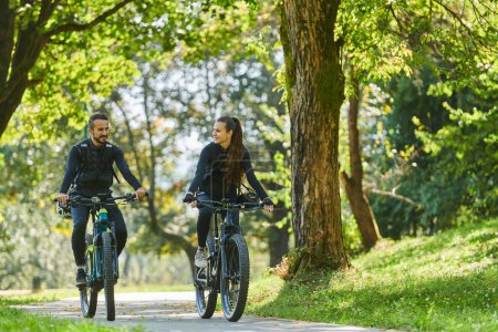Foto de Una pareja feliz, adornada con equipo de ciclismo profesional, disfruta de un romántico paseo en bicicleta a través de un parque, rodeado de modernas atracciones naturales, irradiando amor y felicidad. - Imagen libre de derechos