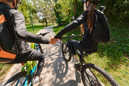 Foto de Una dulce pareja, adornada con ropa de ciclismo, monta sus bicicletas, sus manos entrelazadas en un abrazo romántico, capturando la esencia del amor, la aventura y la alegría en un sendero iluminado por el sol. - Imagen libre de derechos