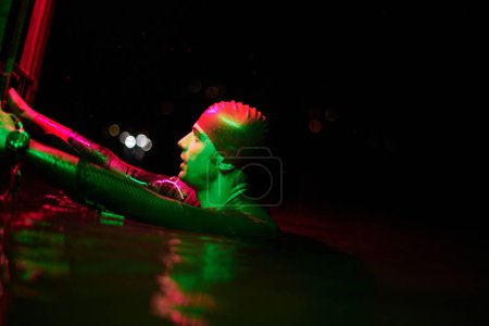 Foto de Nadador triatleta real tener un descanso durante el entrenamiento duro en el lago en la noche oscura luces de color gel de neón - Imagen libre de derechos
