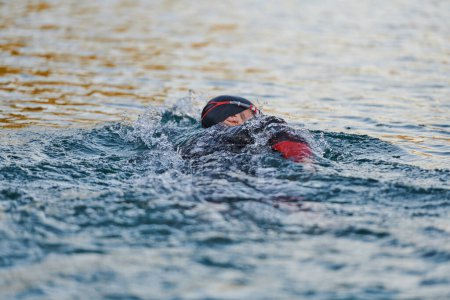 Téléchargez les photos : Athlète de triathlon nageant sur le lac au lever du soleil portant une combinaison. Photo de haute qualité - en image libre de droit