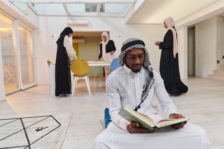 Photo for In the sacred month of Ramadan, an African American Muslim man engrossed in reading the Holy Quran is surrounded by a backdrop where three hijab-wearing women prepare and serve food for iftar - Royalty Free Image