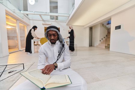 Téléchargez les photos : Au mois sacré du Ramadan, un musulman afro-américain absorbé par la lecture du Saint Coran est entouré d'une toile de fond où trois femmes portant le hijab préparent et servent de la nourriture pour iftar - en image libre de droit