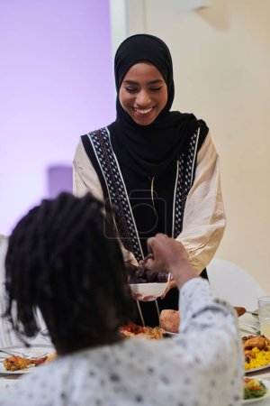 Foto de En una escena conmovedora durante el sagrado mes de Ramadán, una mujer musulmana tradicional ofrece fechas a su familia reunida alrededor de la mesa, ejemplificando el espíritu de unidad, generosidad y cultura. - Imagen libre de derechos