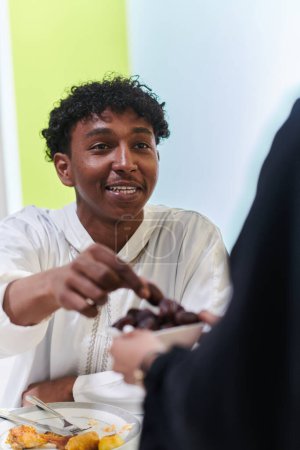 Foto de El hombre musulmán afroamericano delicadamente toma fechas para romper su ayuno durante el mes de Ramadán, sentado a la mesa de la cena familiar, encarnando una escena de reflexión espiritual, tradición cultural y el - Imagen libre de derechos