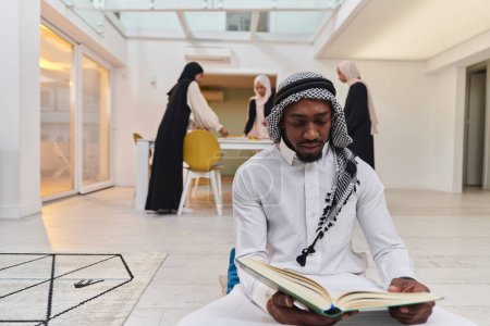 Photo for In the sacred month of Ramadan, an African American Muslim man engrossed in reading the Holy Quran is surrounded by a backdrop where three hijab-wearing women prepare and serve food for iftar - Royalty Free Image