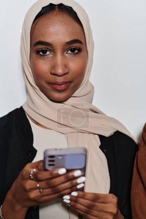 Foto de Una mujer árabe elegante, adornada con un hiyab, se involucra con la modernidad mientras usa un teléfono inteligente, la yuxtaposición de la vestimenta tradicional contra la tecnología contemporánea capturada en el entorno aislado - Imagen libre de derechos