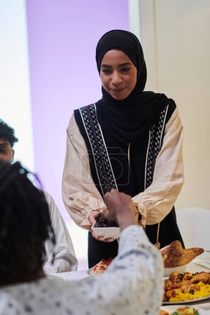 Téléchargez les photos : Dans une scène chaleureuse pendant le mois sacré du Ramadan, une femme musulmane traditionnelle offre des dates à sa famille réunie autour de la table, illustrant l'esprit d'unité, de générosité et de culture. - en image libre de droit