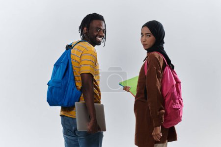 Téléchargez les photos : Étudiante afro-américaine collabore avec sa collègue musulmane, qui travaille avec diligence sur son ordinateur portable, symbolisant un mélange de diversité, d'apprentissage moderne et d'esprit de coopération contre un blanc serein - en image libre de droit