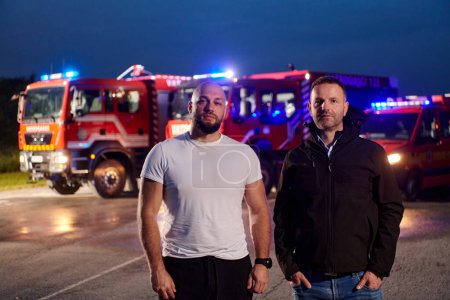Téléchargez les photos : Le groupe de pompiers, vêtus de vêtements civils, se tient devant des camions de pompiers pendant la nuit, mettant en valeur un moment de camaraderie et d'unité au sein de l'équipe alors qu'ils réfléchissent à leurs fonctions et à la - en image libre de droit