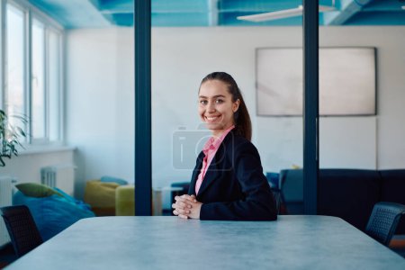 Photo for Successful young female leader in a suit with a pink shirt sitting in a modern glass office with a determined smile - Royalty Free Image