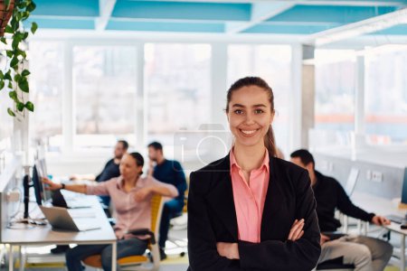 Téléchargez les photos : Portrait de jeune femme d'affaires souriante dans un bureau de démarrage créatif de coworking en espace ouvert. Femme d'affaires prospère debout dans le bureau avec copyspace. Coworkers travaillant en arrière-plan. - en image libre de droit