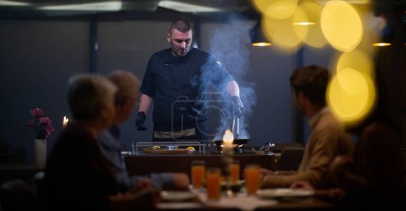 Téléchargez les photos : Dans un restaurant, un chef professionnel présente un steak pétillant cuit sur une flamme nue, tandis qu'une famille musulmane européenne attend avec impatience leur repas à l'iftar pendant le mois sacré du Ramadan - en image libre de droit