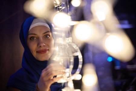 In a modern restaurant ambiance, a woman in a hijab captures a selfie beside glowing lights, showcasing contemporary style and cultural diversity in a trendy urban setting
