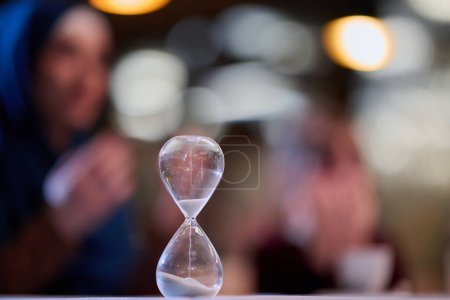 A symbolic hourglass counts down the time to iftar meal, while in the background, an Islamic family devoutly prays, capturing a poignant moment of spiritual connection and anticipation during Ramadan.