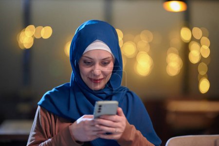 Téléchargez les photos : Une belle musulmane européenne, ornée d'un hijab, utilise son smartphone pour se préparer rapidement à l'iftar pendant le mois sacré du Ramadan, incarnant le mélange de tradition et de modernité en elle - en image libre de droit