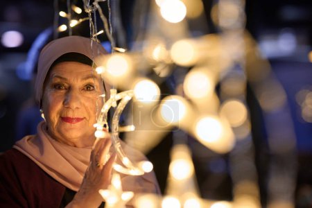 In a modern restaurant ambiance, a woman in a hijab captures a selfie beside glowing lights, showcasing contemporary style and cultural diversity in a trendy urban setting. 