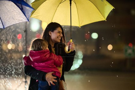 Eine Mutter trägt ihre kleine Tochter zärtlich bei sich, während sie sie in einer regnerischen Nacht mit einem Regenschirm abschirmt. Sie verkörpert die schützende Liebe und Wärme der mütterlichen Fürsorge inmitten des Stadtbildes..