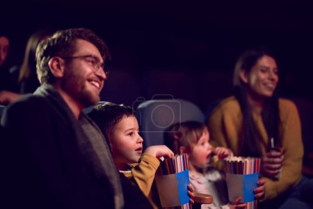 A modern family enjoys quality time together at the cinema, indulging in popcorn while watching a movie with their children.