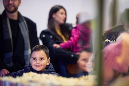 Téléchargez les photos : Un jeune couple avec leurs enfants se tient devant le cinéma, achetant du pop-corn fraîchement éclaté avant le début du film et entrant dans le cinéma. - en image libre de droit