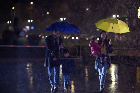 Foto de En medio de una noche urbana lluviosa, una feliz pareja lleva a sus hijos a dar un paseo por las calles de la ciudad, dirigiéndose hacia el cine para una deliciosa salida de películas familiares. - Imagen libre de derechos
