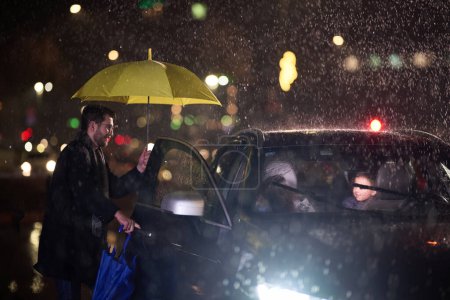 Un hombre de familia abre amorosamente el coche para su familia en tiempo lluvioso, protegiéndolos con un paraguas.