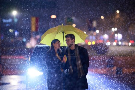 Téléchargez les photos : Dans l'ambiance romantique d'une nuit pluvieuse, un couple heureux se promène dans la ville, partageant des moments tendres sous un parapluie jaune, entouré par la lueur scintillante des lumières urbaines. - en image libre de droit