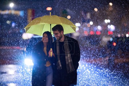 Foto de En el ambiente romántico de una noche lluviosa, una feliz pareja camina por la ciudad, compartiendo momentos tiernos bajo un paraguas amarillo, rodeado por el resplandor brillante de las luces urbanas. - Imagen libre de derechos
