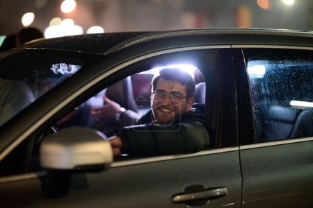 A modern businessman, donning eyeglasses, confidently drives his sleek car through the urban cityscape on a rainy night, epitomizing sophistication and success in his professional endeavors.