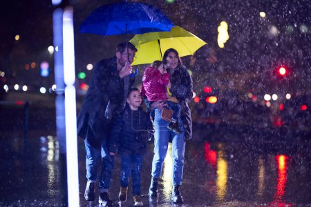 Photo for In the midst of a rainy urban night, a happy couple takes their children on a stroll through the city streets, heading towards the cinema for a delightful family movie outing. - Royalty Free Image