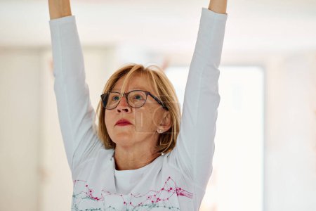 Téléchargez les photos : Dans un espace ensoleillé, une femme âgée pratique gracieusement le yoga rajeunissant, se concentrant sur les étirements du cou, du dos et des jambes, incarnant la sérénité et le bien-être.. - en image libre de droit