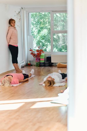 Photo for A skilled trainer oversees a group of senior women practicing various yoga exercises, including neck, back, and leg stretches, in a sunlit space, promoting wellness and harmony. - Royalty Free Image