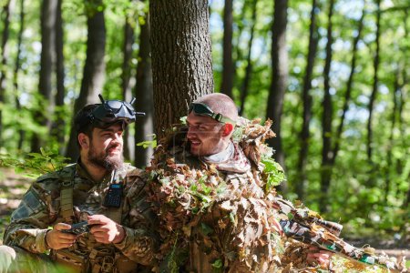Téléchargez les photos : Un tireur d'élite habile et un soldat opérant un drone avec des lunettes VR élaborent une stratégie et observent l'action militaire tout en étant caché dans la forêt. - en image libre de droit